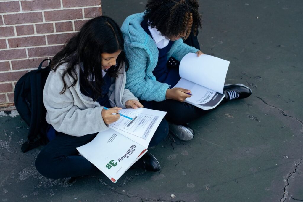 From above of adorable diligent pupils learning and reading textbooks while sitting near brick wall